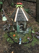 Round, wooden fairy house with shingled roof & moss lawn.